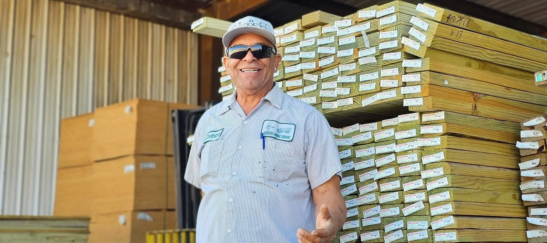 Smiling Sorrento team member in a company cap and shirt in front of stacked lumber at the yard